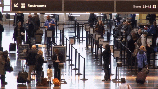 Travelers wait in multiple security screening lines at an airport checkpoint beneath overhead signs for restrooms and gates 35–45.