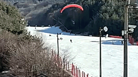 Paraglider with a red canopy flies low over a ski resort slope as skiers descend below and spectators watch from the side.